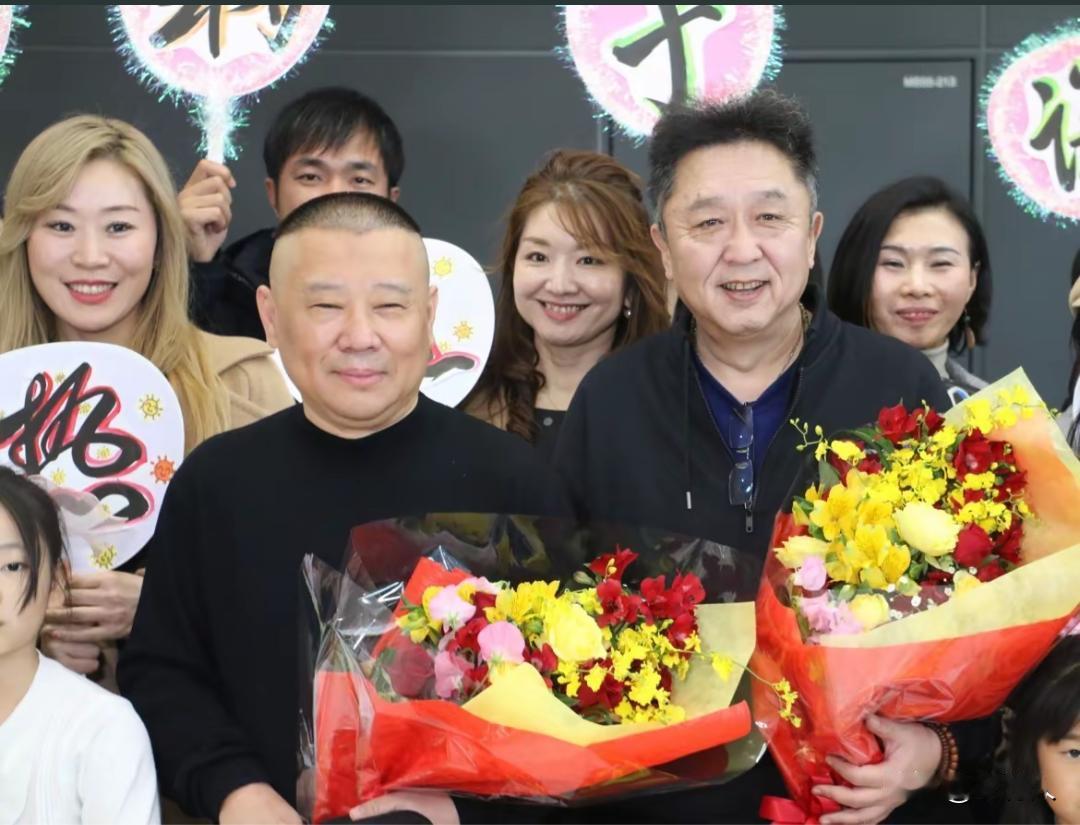 郭德纲和于谦手捧鲜花，身后是剧组工作人员。是线下活动杀青合影现场，整体氛围热闹喜