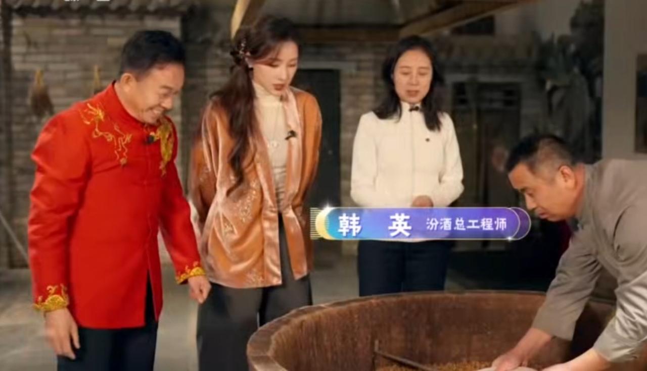 在汾酒酿造工坊里，总工程师韩英与众人一同观摩酿酒工序，匠人俯身操作大缸，尽显传统