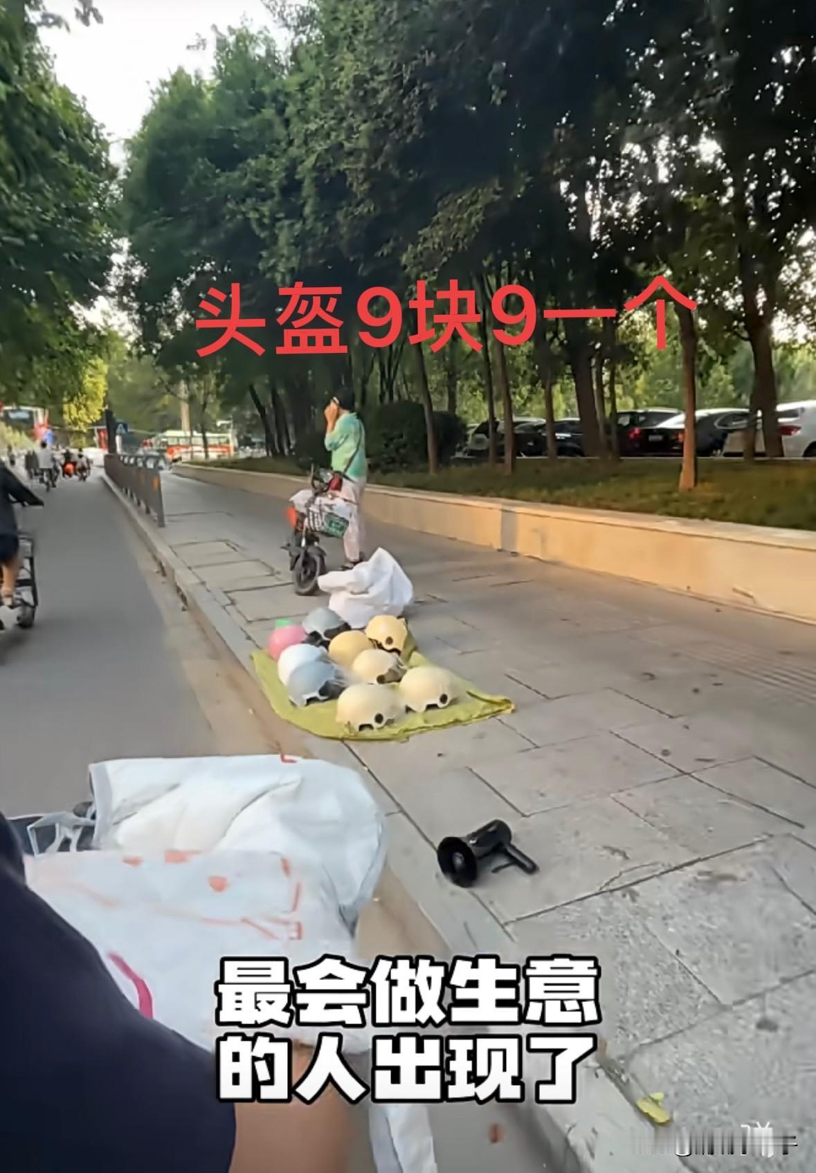 这就是传说中的商业奇才吧。
女子在十字路口不远处摆摊卖头盔，旁边大喇叭反复喊着“