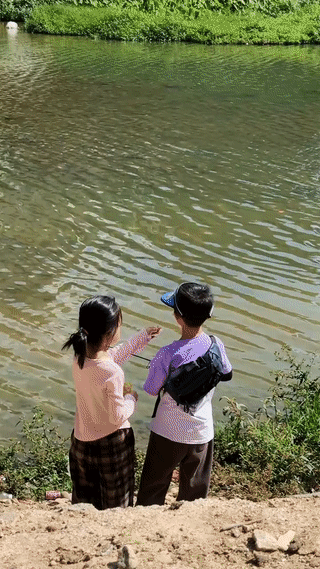 广东汕尾，一名小女孩陪着小男孩站在河边钓鱼。
小女孩还将手里的零食喂给小男孩吃。