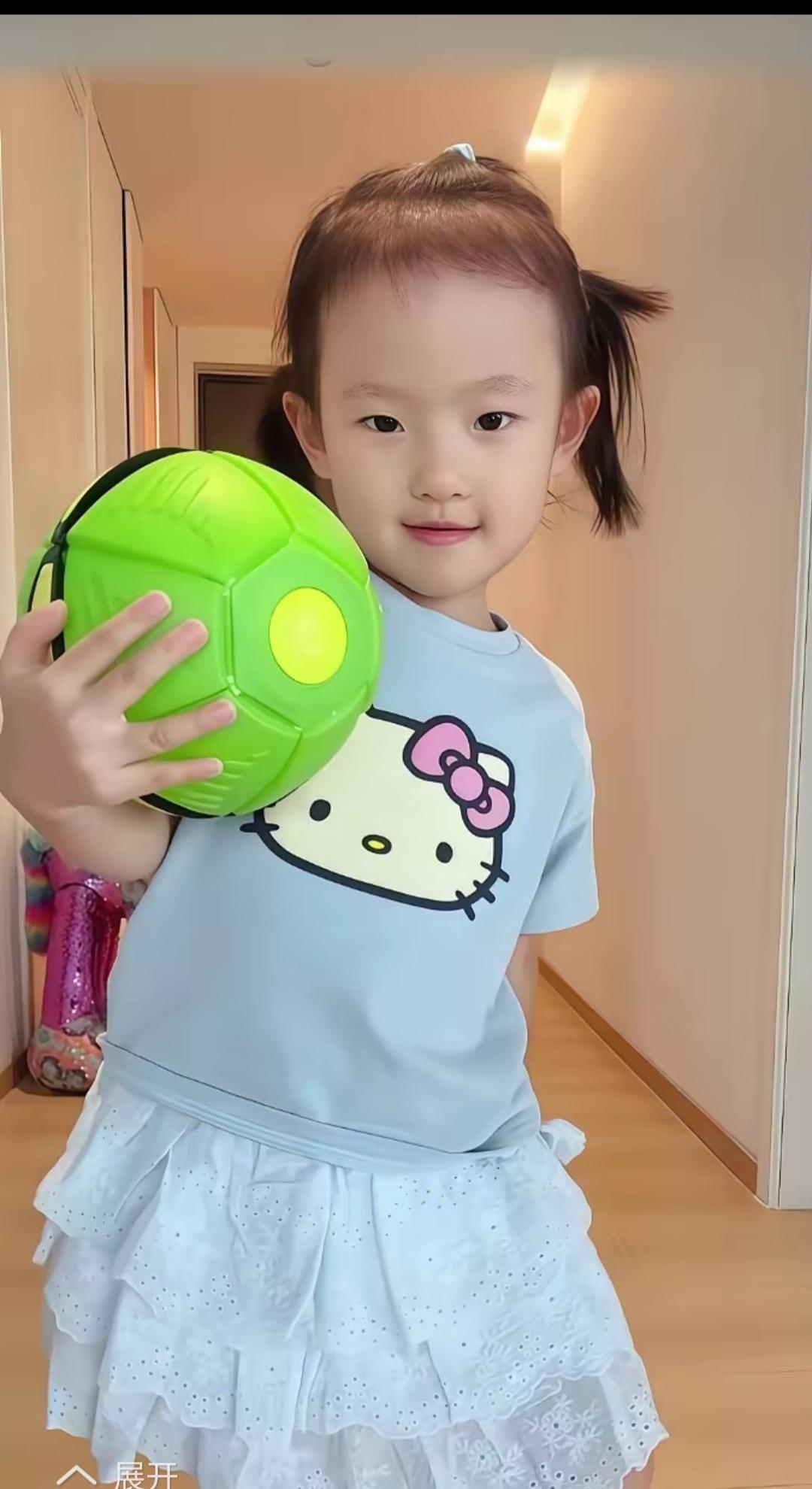 照片中的闪闪穿着公主裙小T恤，眨着可爱的羊角辫，抱着绿色的小球，真是越长越可爱，