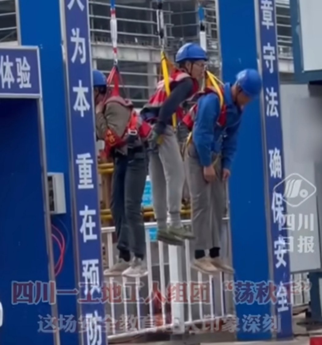 工地把3名工人吊在安全带使用体验区蓝色安全帽，戴眼镜，一般应该是技术人员或者管理