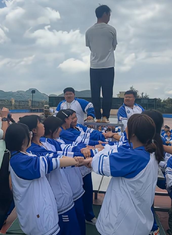 十名女同学挽着手当垫子，男生从高处往后倒从缝隙中跌落。
网友说，不用眼睛也能瞄准
