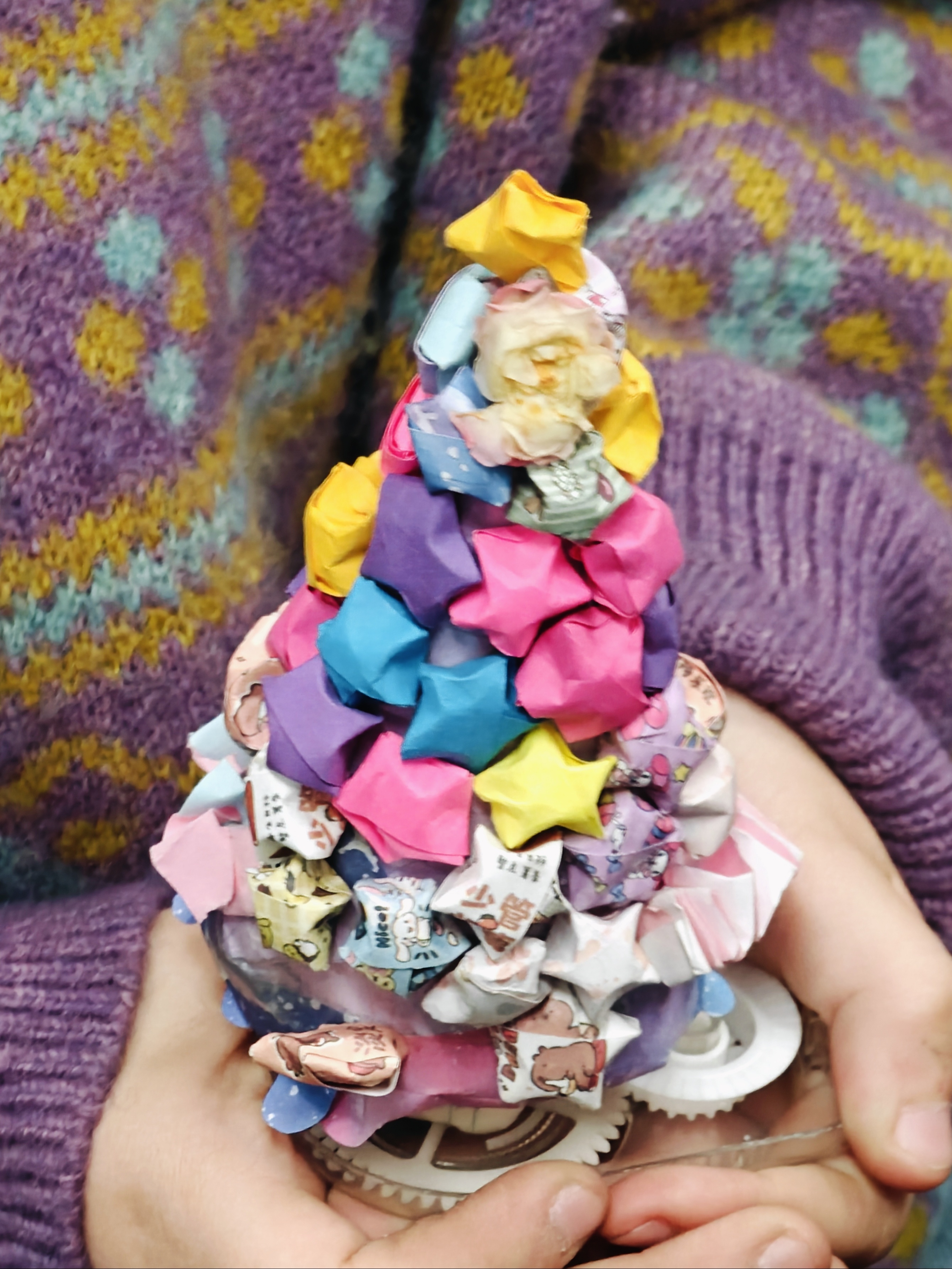 学生M用零食包装和修正带外壳做的圣诞树。她用一种照顾受伤小鸟的手势把树含在掌心，