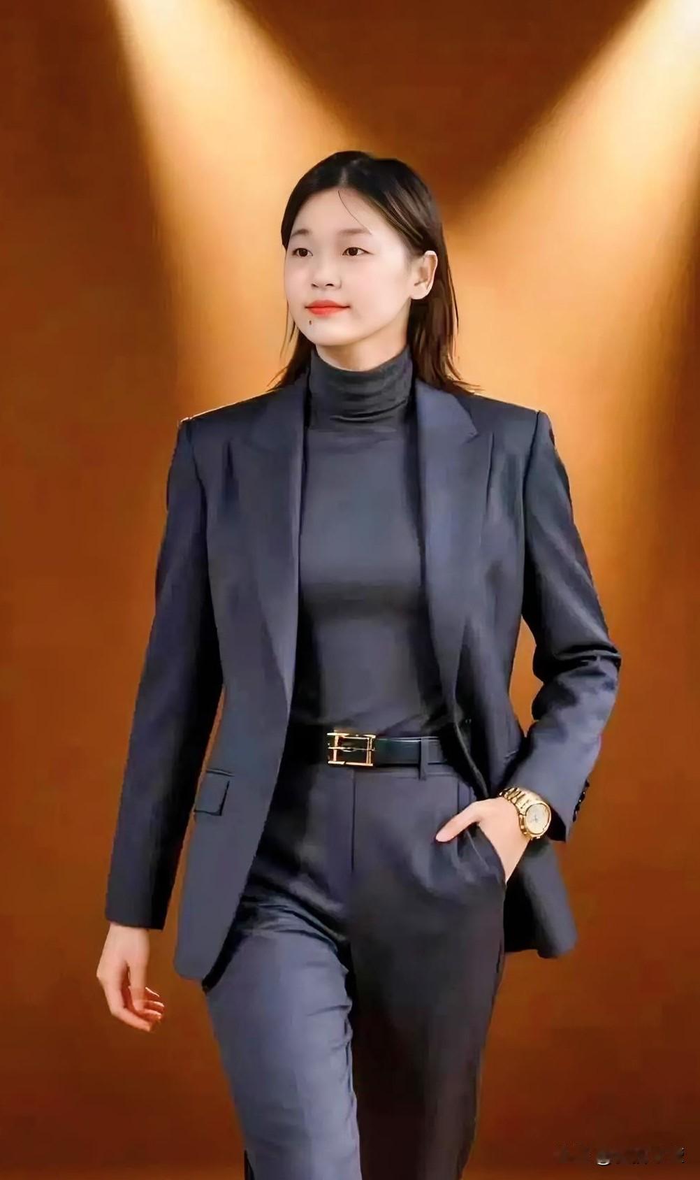 她身着深灰色高领内搭与同色系西装套装，搭配金色腰带与腕表，利落又精致。灯光勾勒出