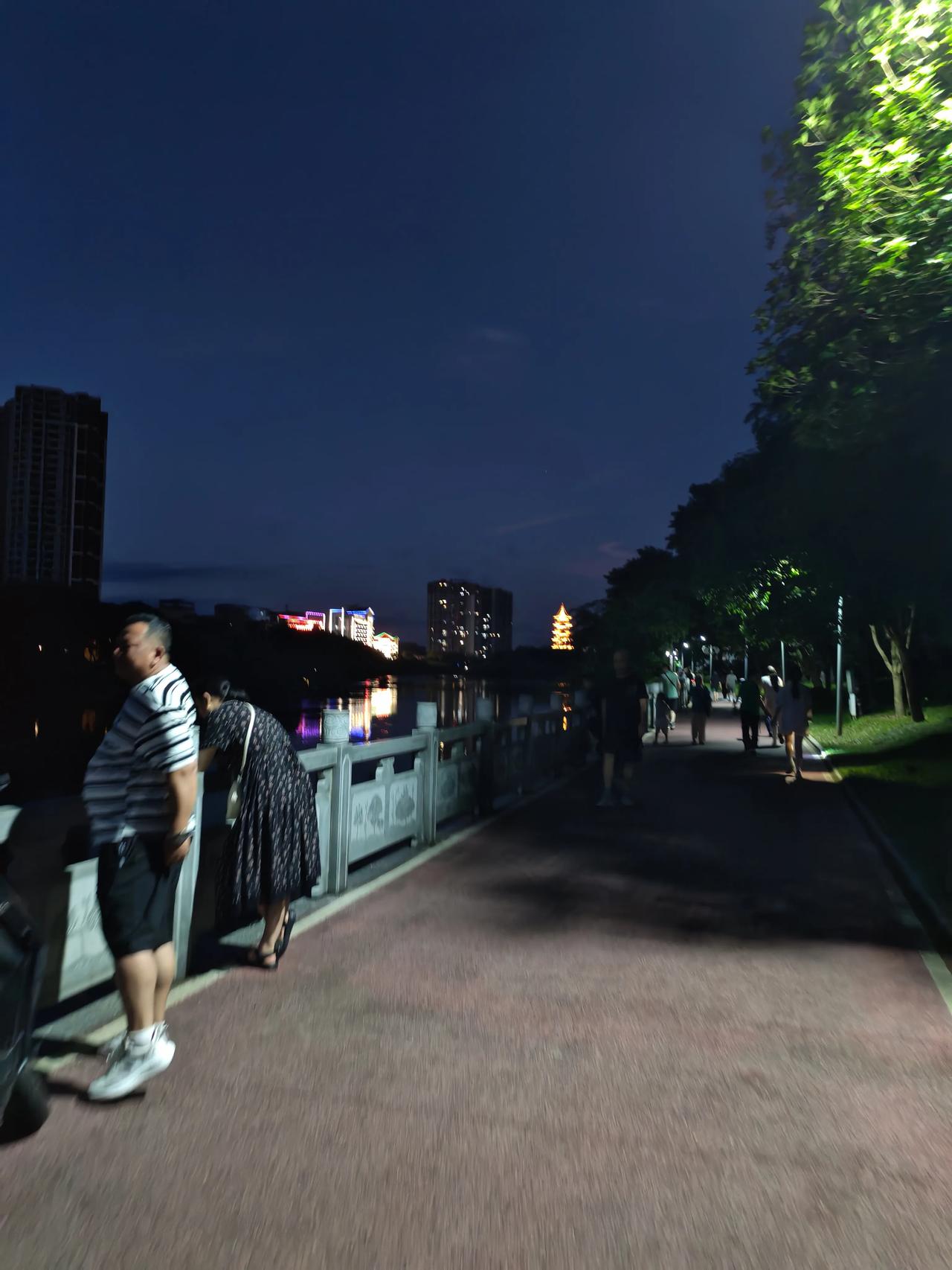 夜色渐浓，漫步滨河公园的慢行步道。绿树成荫，灯光点缀林间，远处古塔金光熠熠，高楼