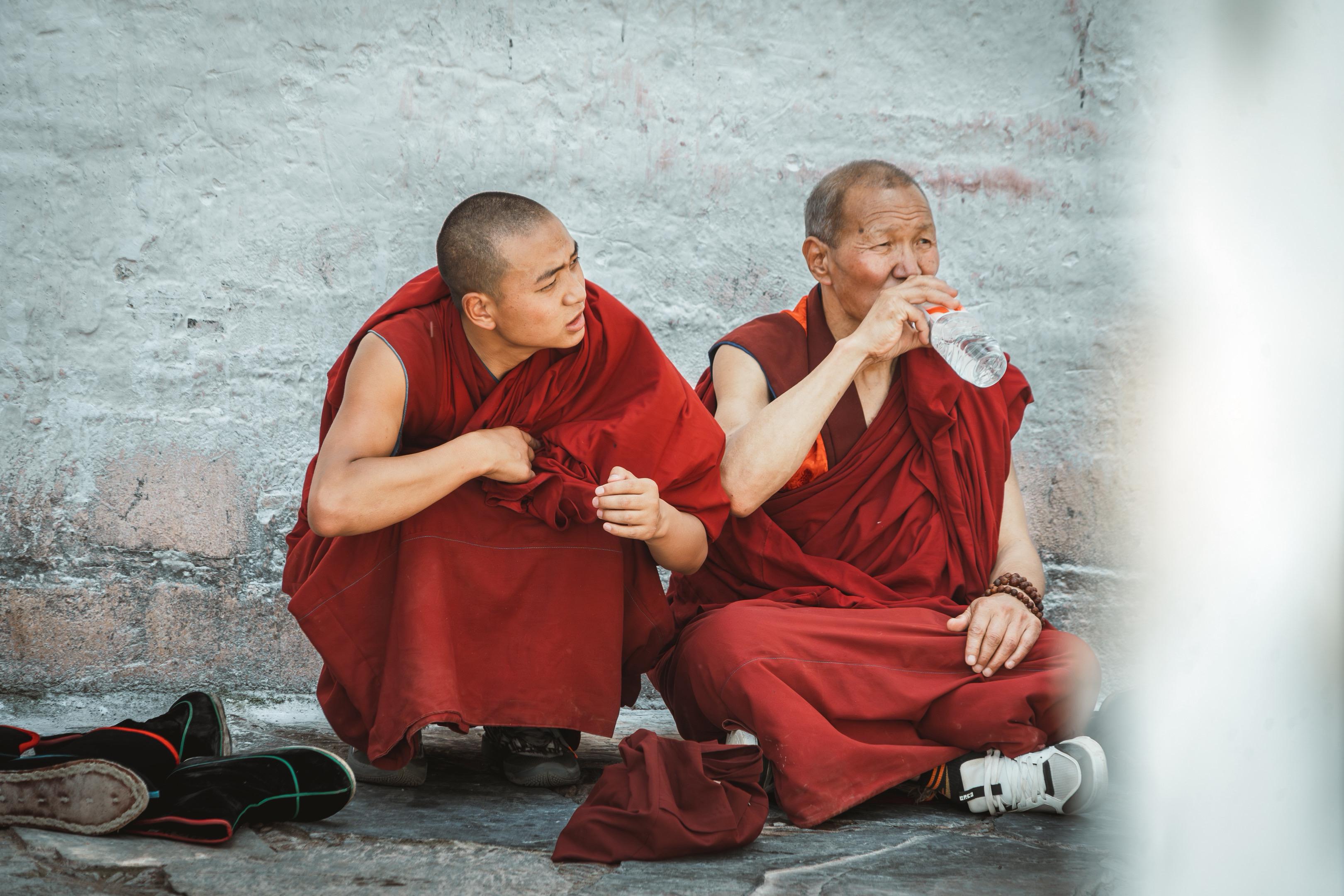 在拉卜楞寺的白墙下，两位僧人席地而坐。年轻僧人侧身似在交谈，年长僧人手...
