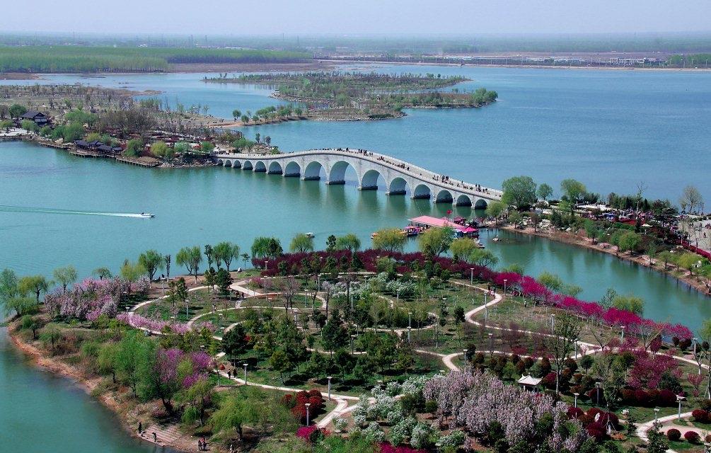 画面中，西双湖的湖水澄澈如镜，一座多拱石桥横跨湖面，连接起湖岸与湖心岛。桃花岛上