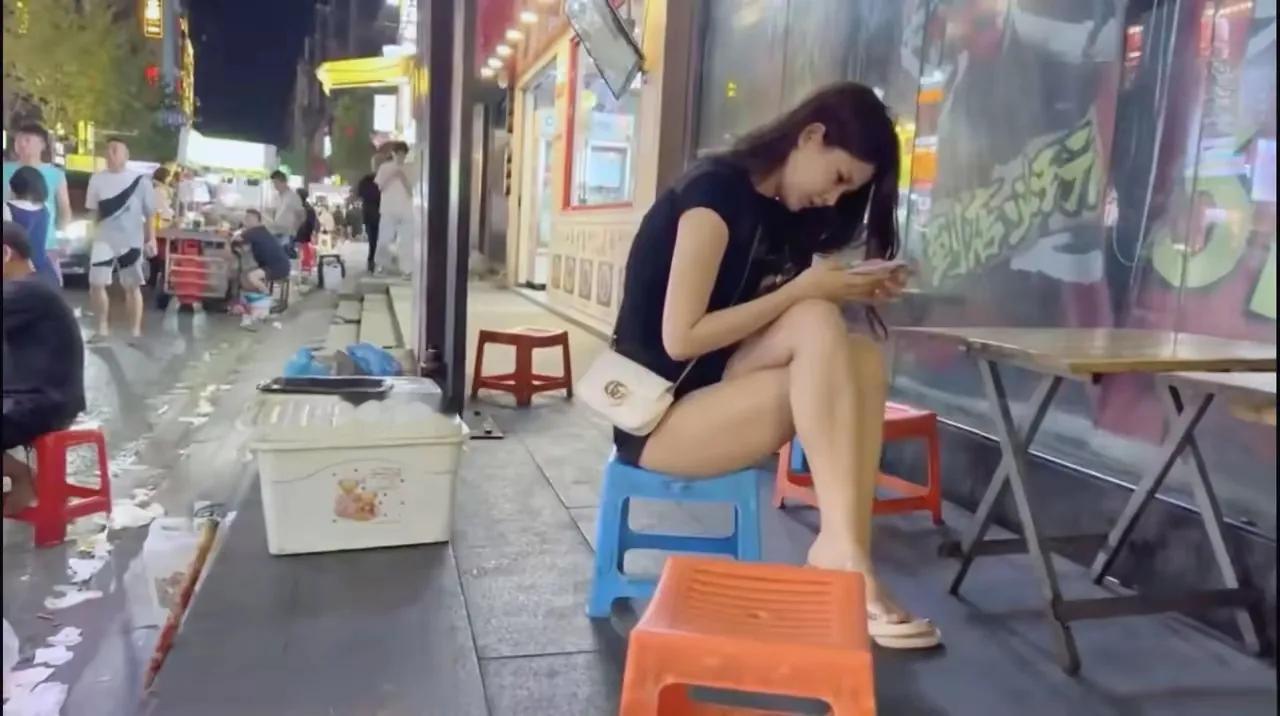 夜幕低垂，路边摊灯火阑珊，一位绝色佳人静坐其间，手机屏幕映出她孤独的轮廓。突然间
