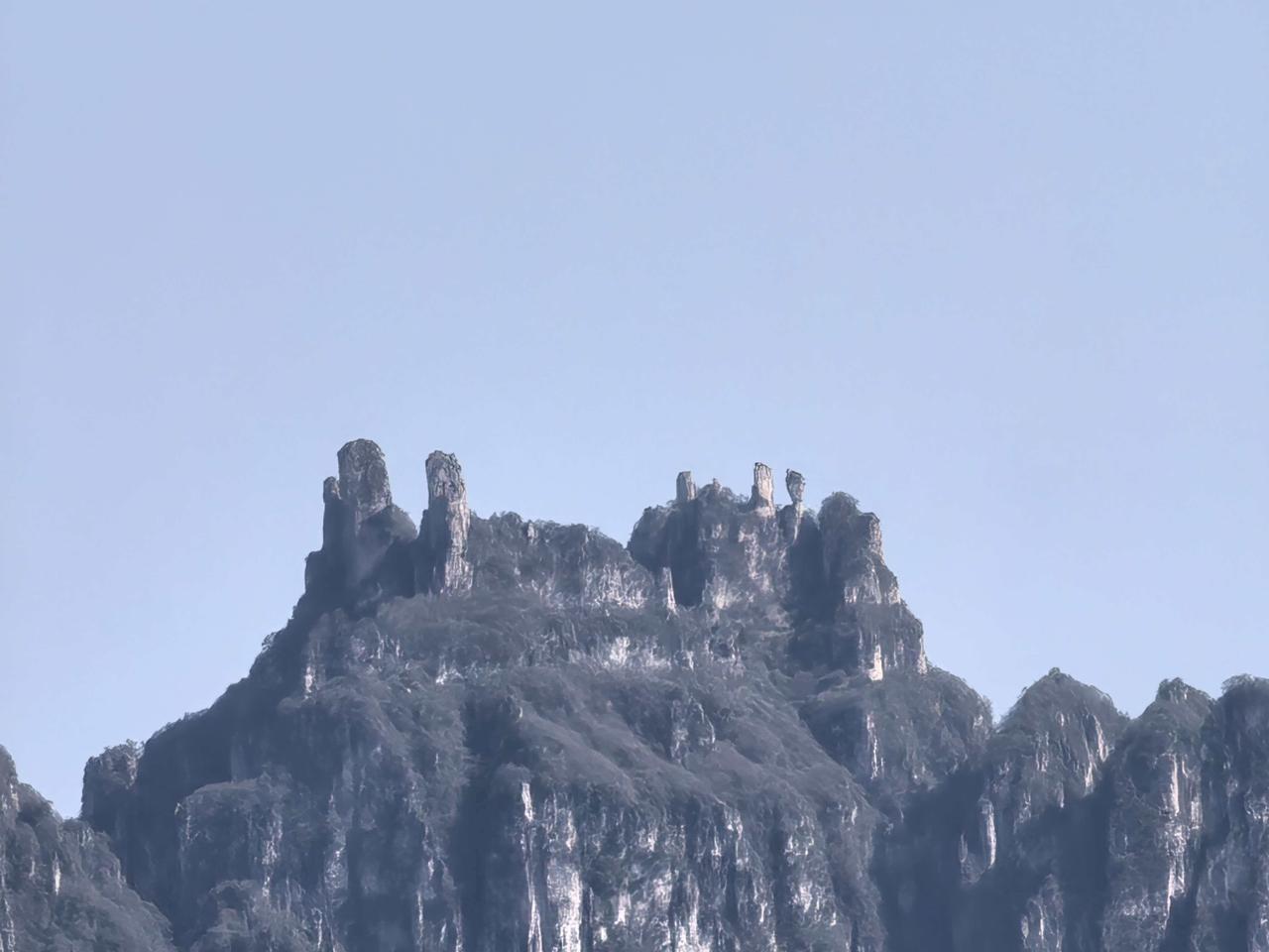 晒山里的景色澄澈浅蓝的天幕铺展在画面上方，几座嶙峋陡峭的石峰刺破山峦，挺拔的岩柱