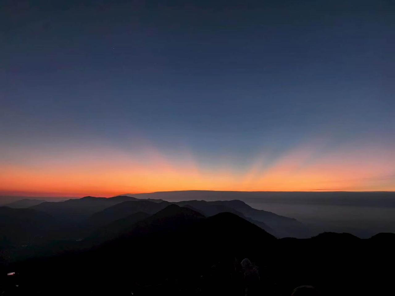夜色还沉在群山褶皱里，远山化作深浅错落的剪影。天际线慢慢晕开橘红霞光，温柔的光带