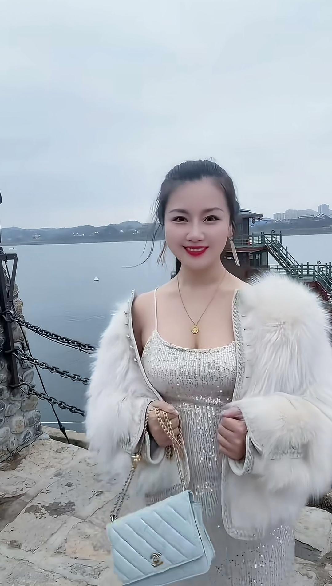 这位美女在湖边拍照，穿着闪亮的衣服和毛茸茸的外套。