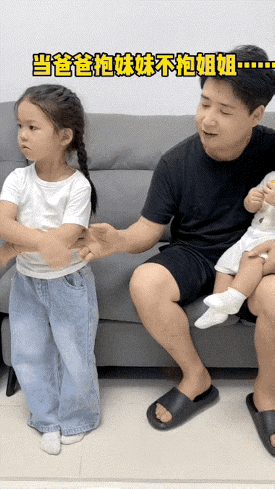 姐姐抱着肩膀在沙发旁站着，爸爸抱着妹妹用手来拉姐姐的胳膊，姐姐胳膊一甩“别碰我”