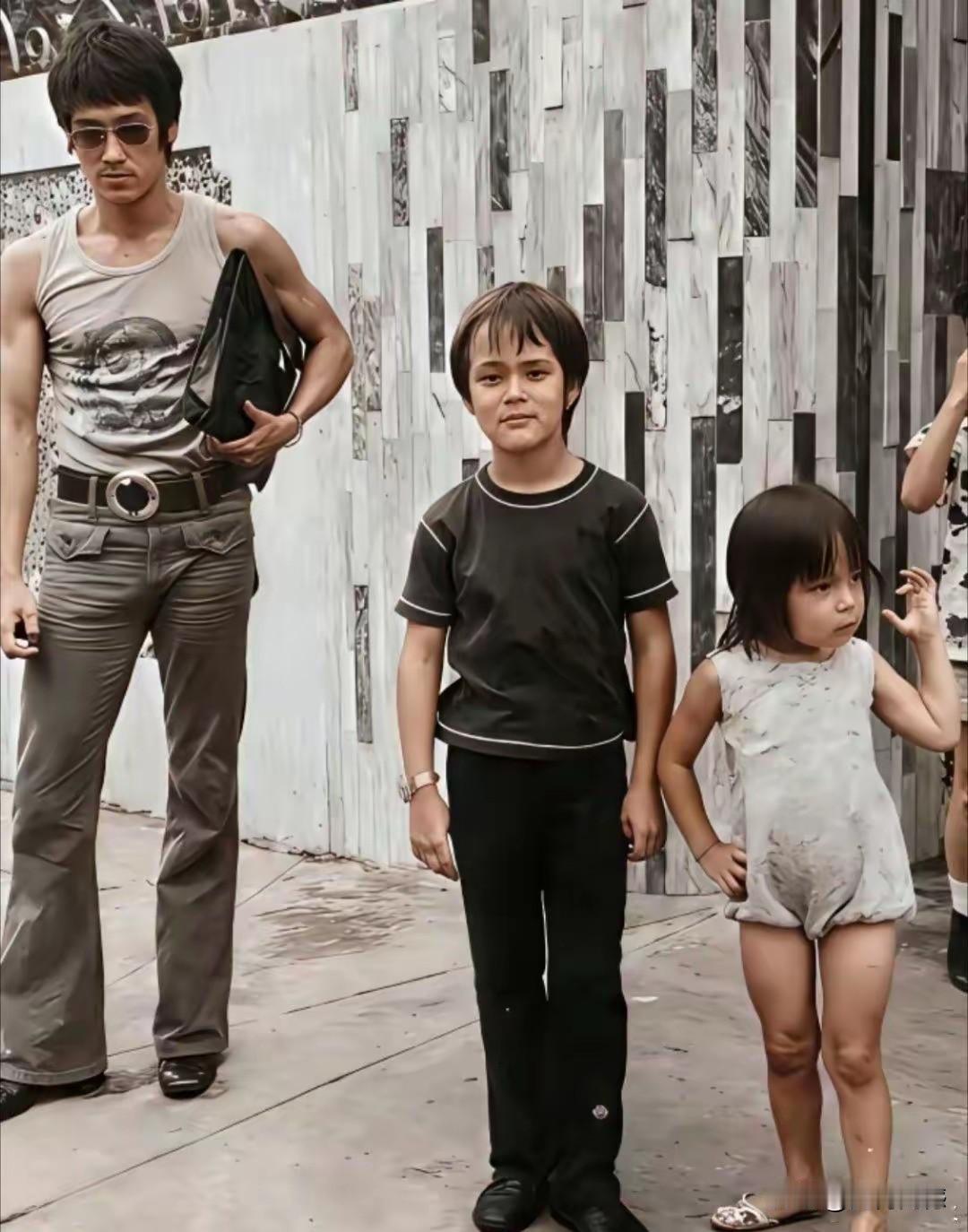 照片中的李小龙，夹着皮包，戴着墨镜，看起来就像是一个硬汉，站在他身边的这两个孩子