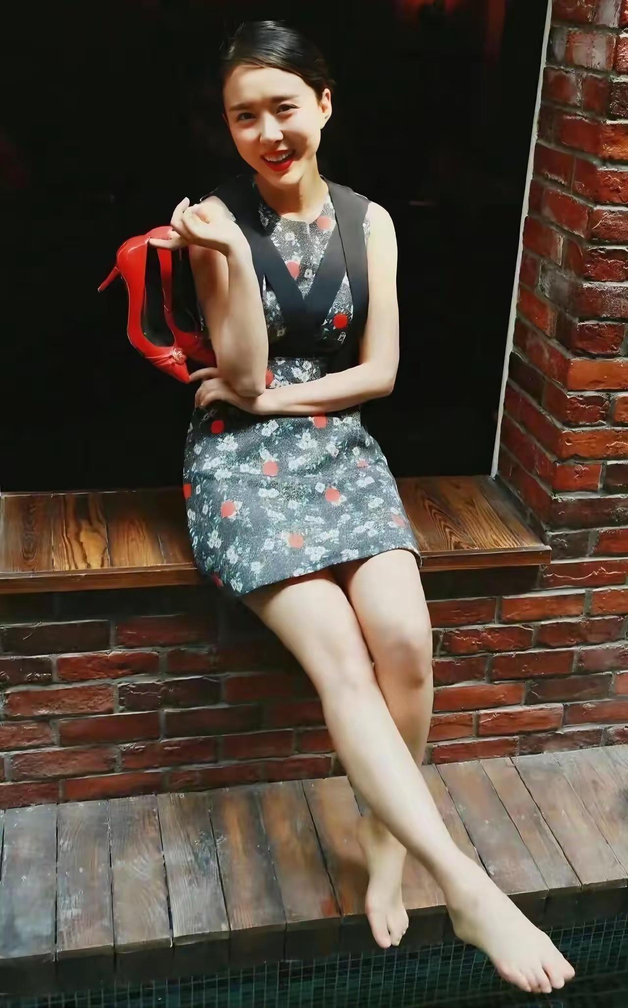这位小姐姐坐在砖墙上，脚翘着，手里拿着高跟鞋。