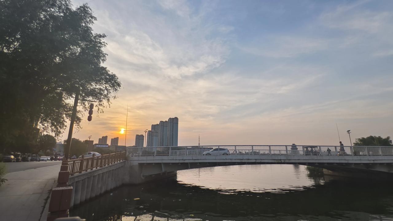 石桥横跨流水，桥面车来人往，行人步履悠然。河水静静荡漾，完整复刻出落日与桥身的轮
