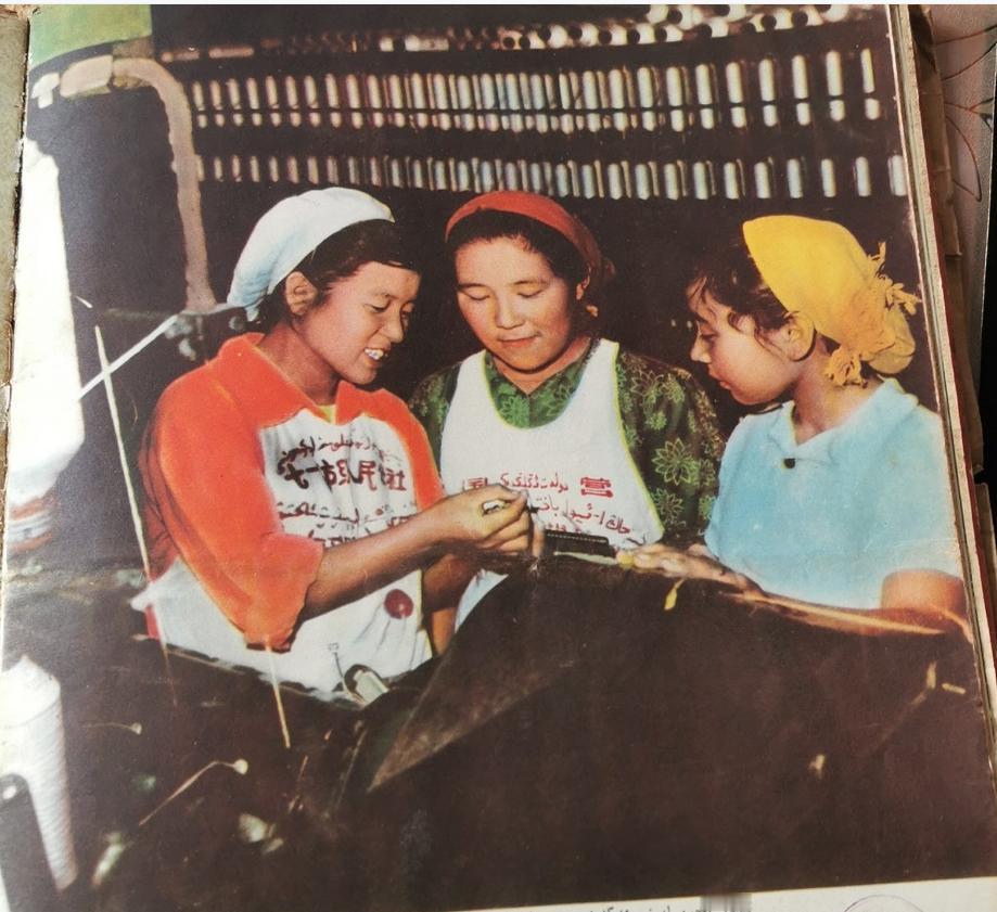 照片展现了三位身着工装、头裹头巾的维吾尔族纺织女工，在车间内交流技艺的场景。她们