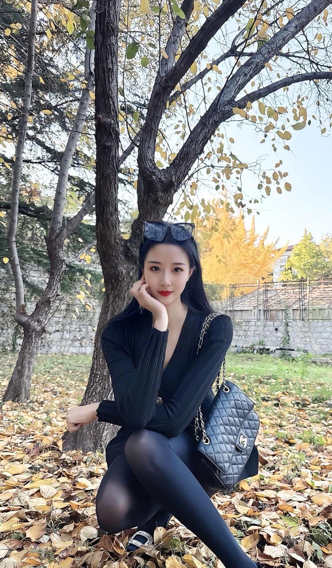 秋日暖阳下，黑衣美人倚树而立，落叶纷飞，时尚与自然完美融合。