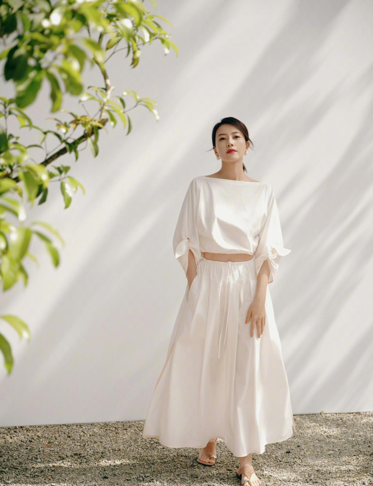 身着一套简约的白色套装，上衣为露腰短款设计，搭配同色系的宽松长裙，脚踩一双简约的