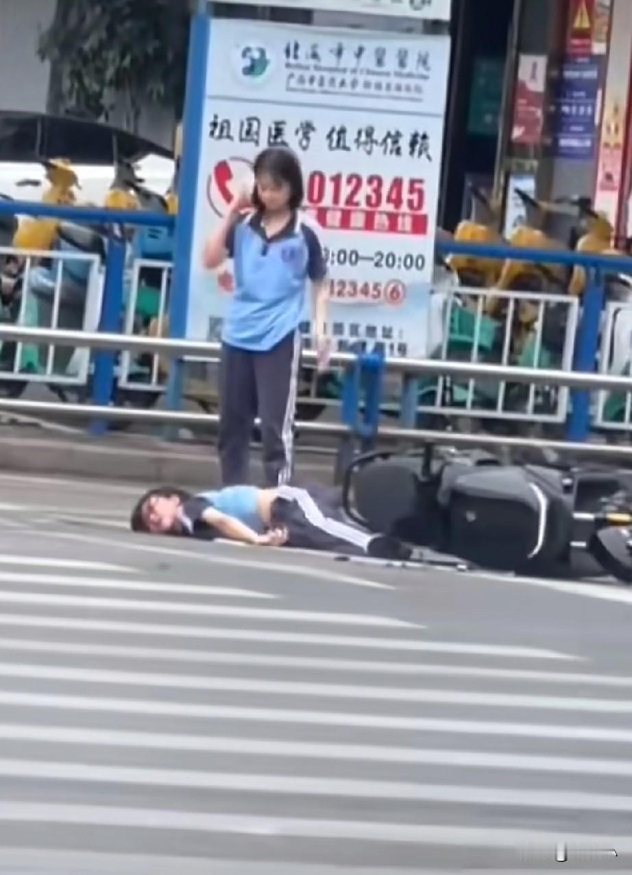 两个女孩骑车摔倒，其中一个倒在地上不停的抽搐着，另一个一边看着同伴，一边打电话，
