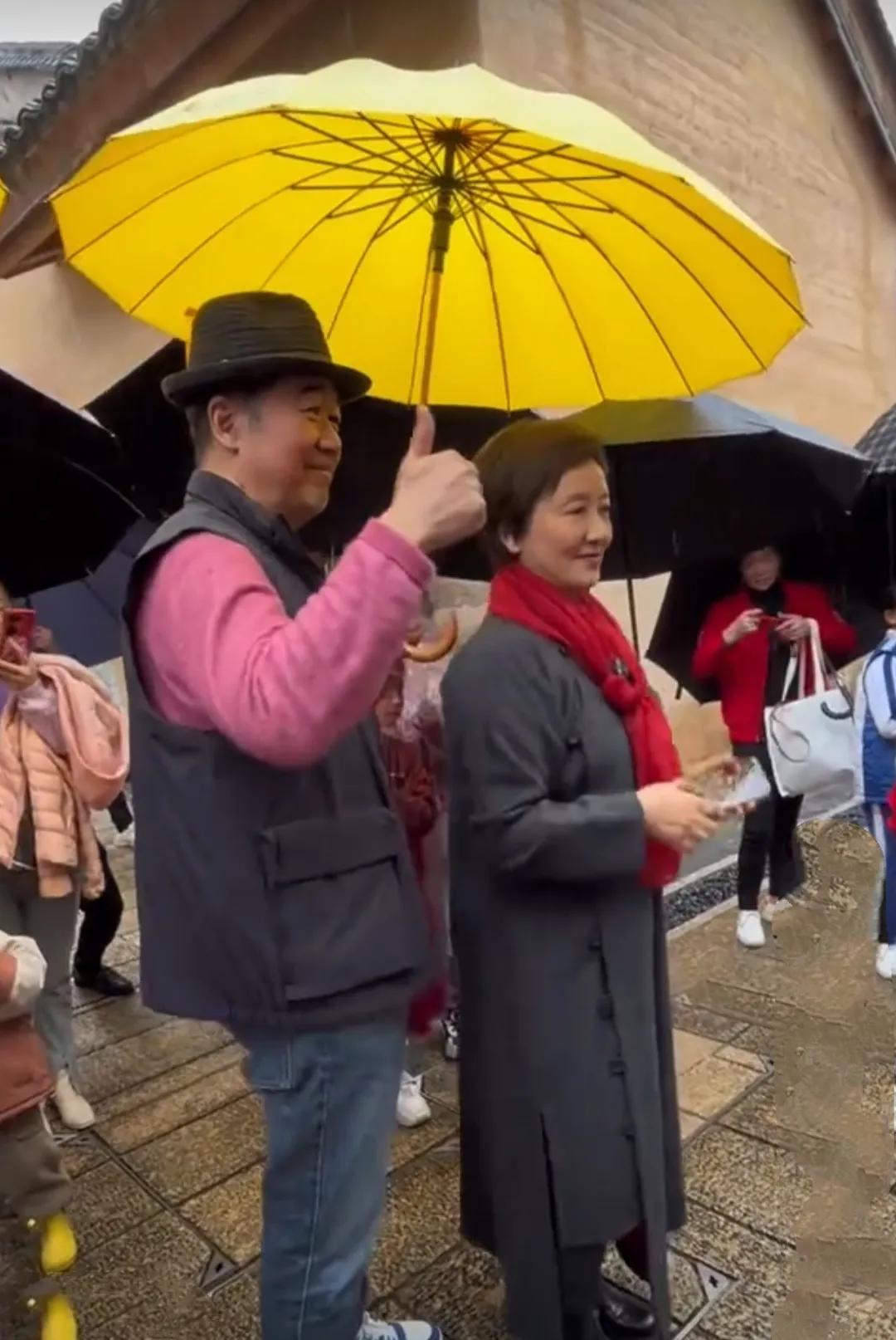 张国立邓婕夫妇雨中逛街，打着黄色雨伞，张国立戴着黑色礼帽，灰黑色羽绒马甲，粉色毛