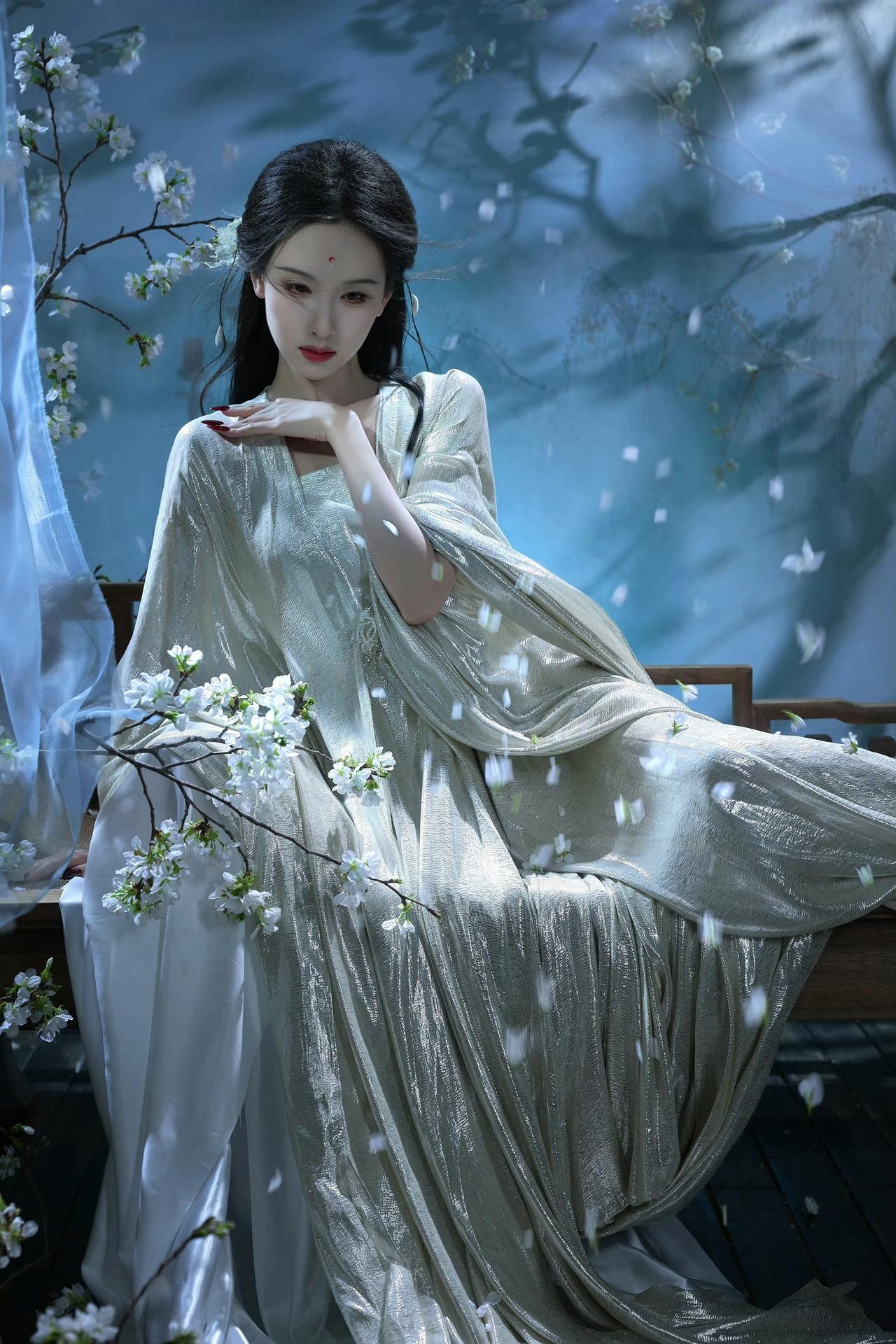 她身着一袭带有光泽感的浅色古风长裙，置身于带有白色花枝、飘着花瓣的朦胧场景中，造
