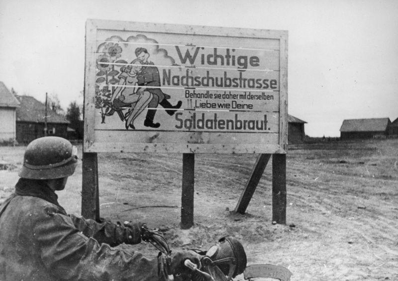一名德军摩托车兵停靠在路边的德军宣传牌旁。
宣传牌上的文字写道：“这是重要的补给