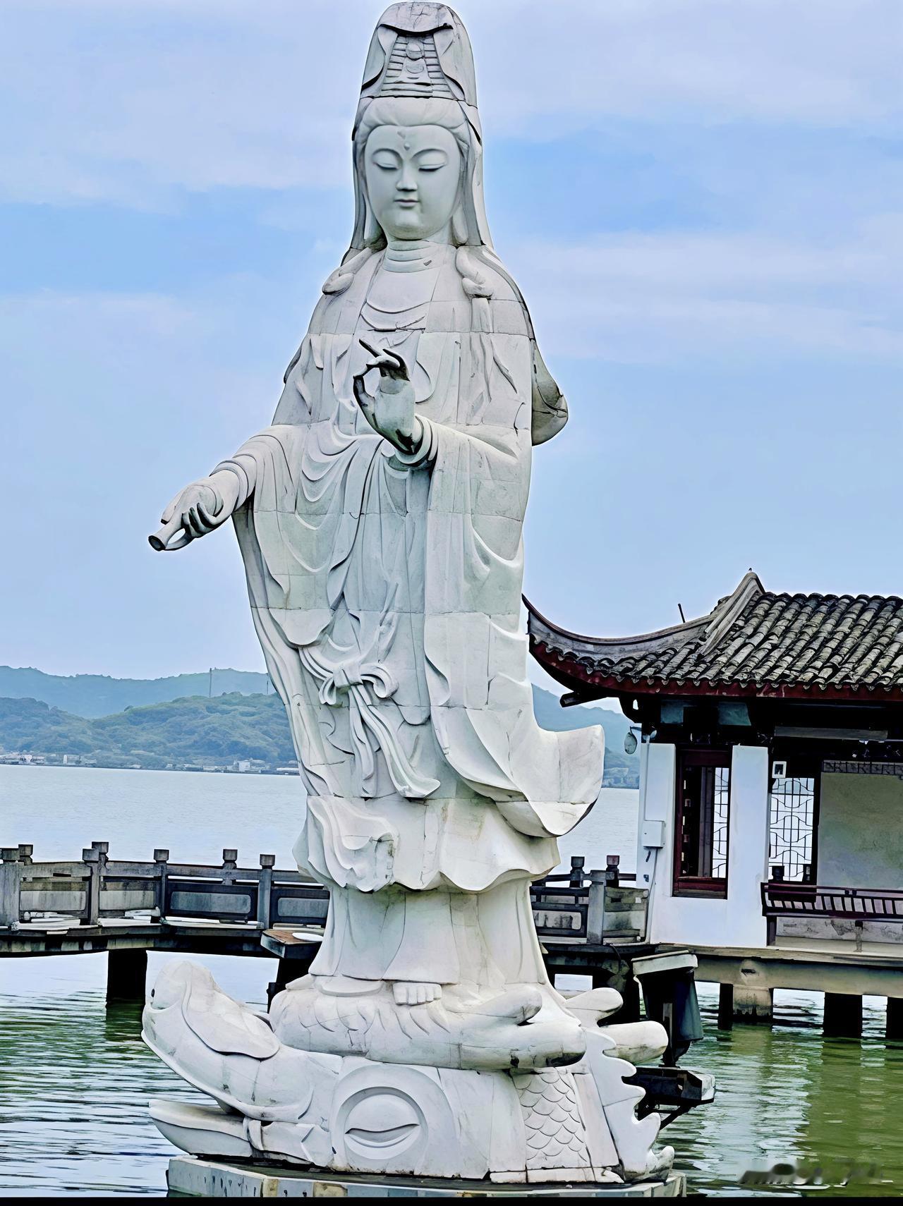 宁波东钱湖之上，白玉观音石像临水而立，衣袂翩然似随湖风轻拂。脚下碧波荡漾，佛影倒