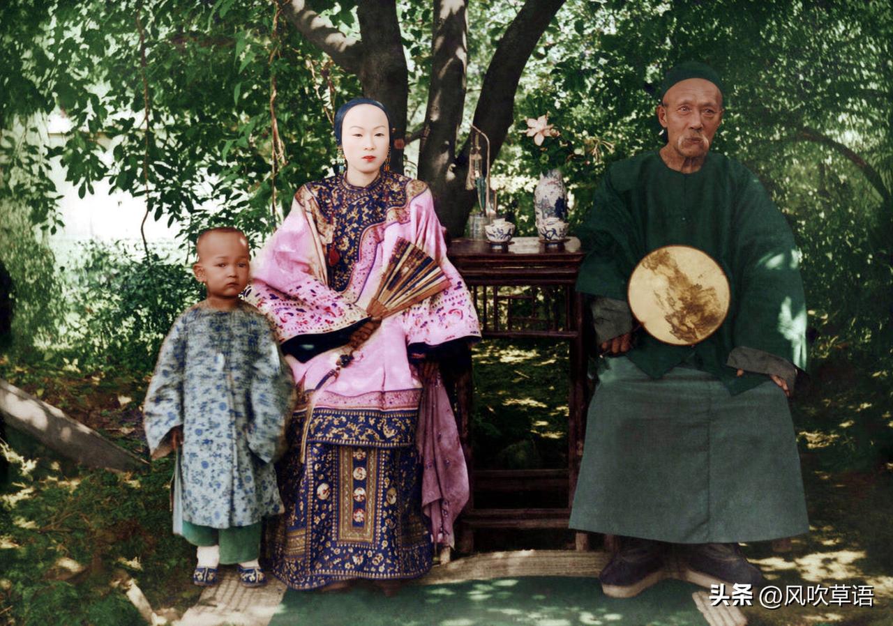 1871 年，一对老夫少妻坐在树荫下的合影，丈夫握团扇，妻子持折扇，旁边站着他们