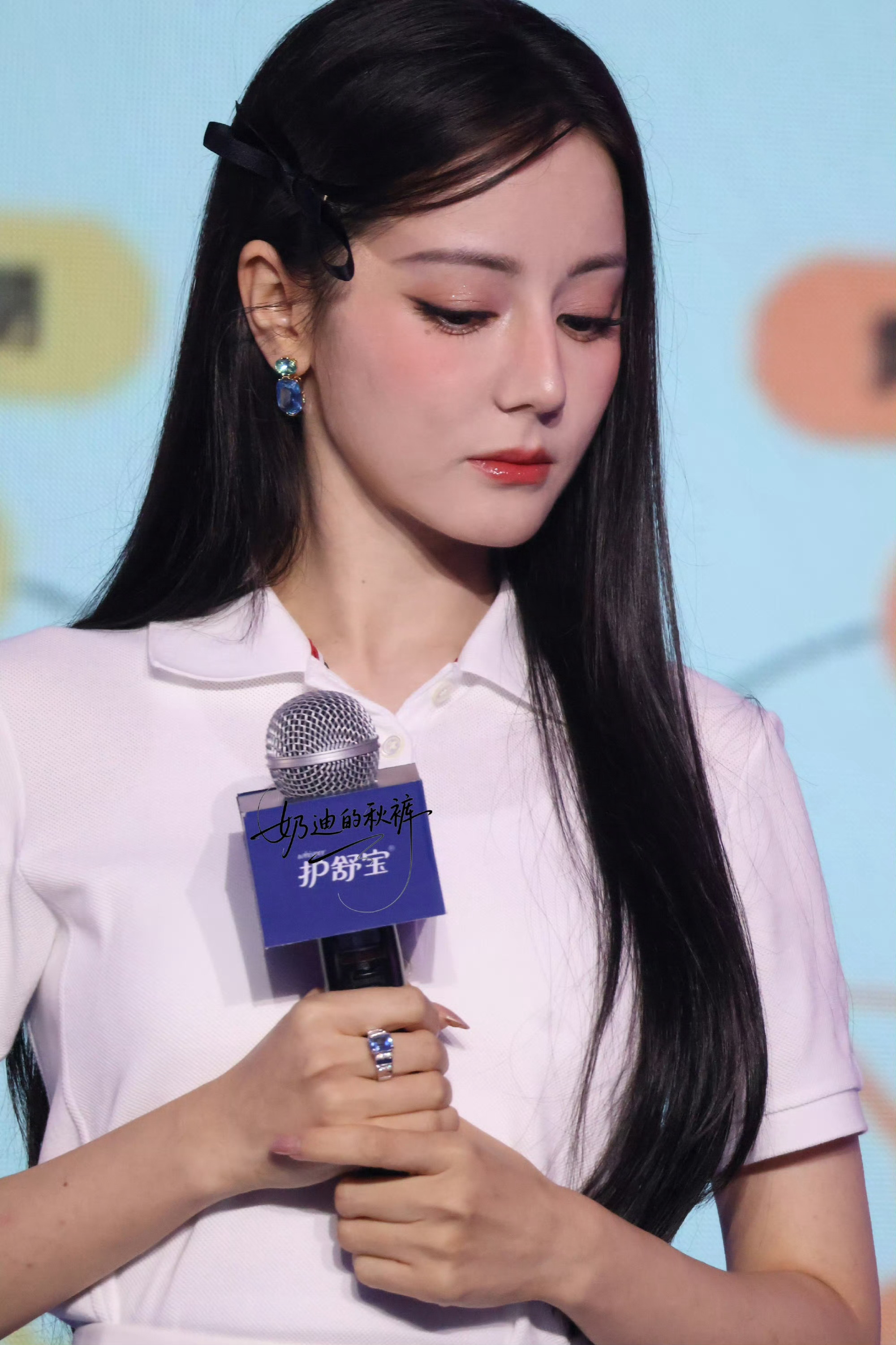 在活动现场，迪丽热巴一袭简约白衫，发丝垂坠，发间黑蝴蝶结添了几分娇俏。手捧护舒宝