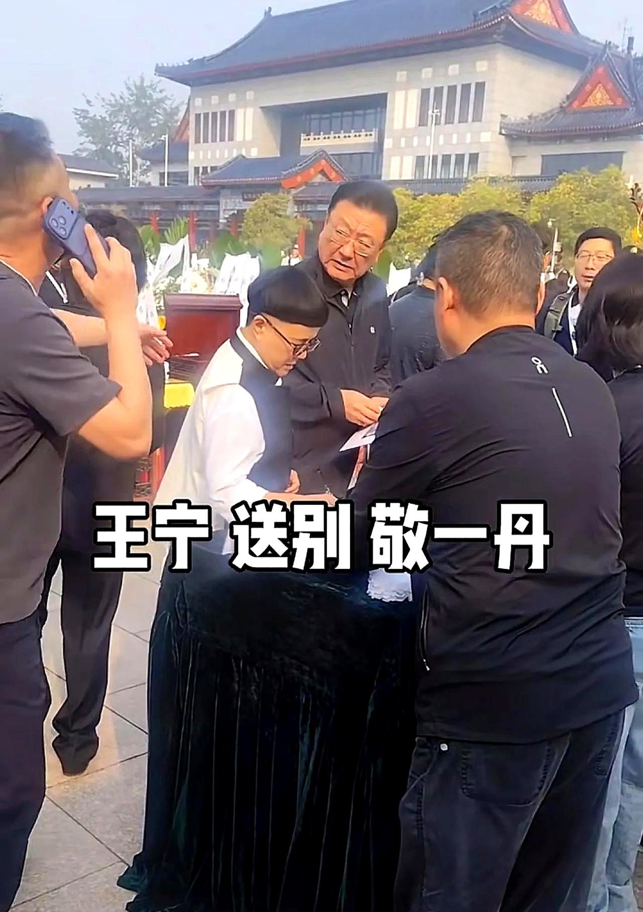 八宝山东礼堂外，风一阵阵吹过花圈，队伍弯到街角。
布置极简，灯光收着，整场只为一