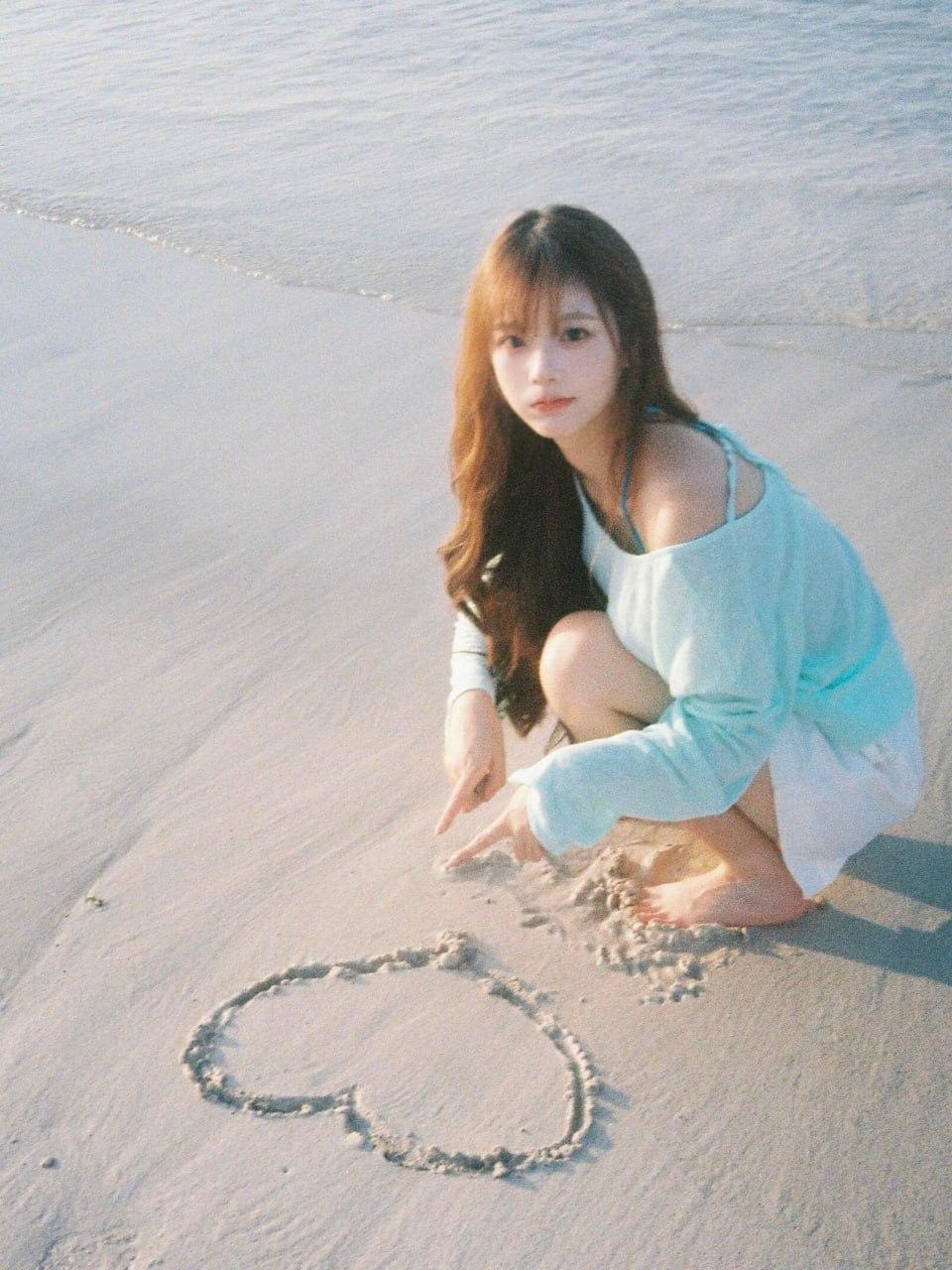 一位长发女孩跪坐在沙滩上，她身着浅蓝色露肩长袖上衣和白色短裤，赤着双脚。女孩伸出