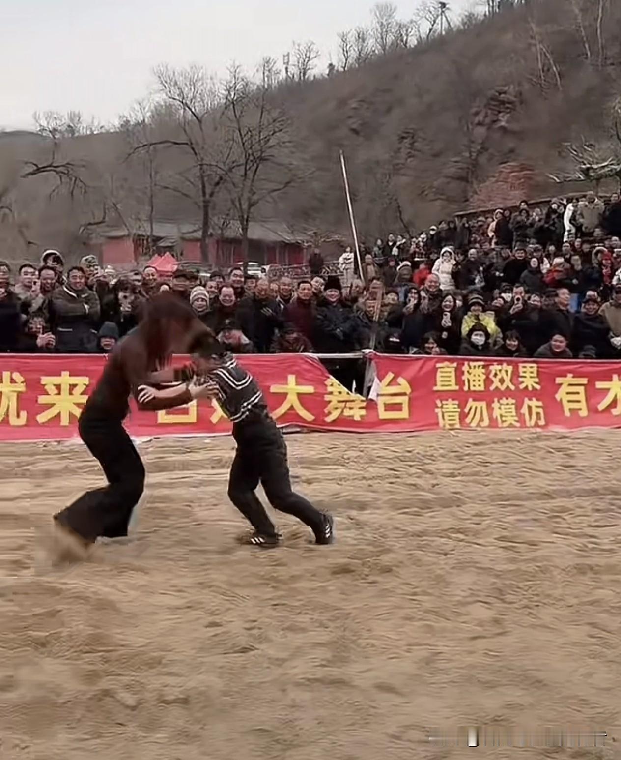 沙地上演着一场别开生面的较量：几位身形矫健的女子相继围着一个矮个子男子嬉戏般摔跤