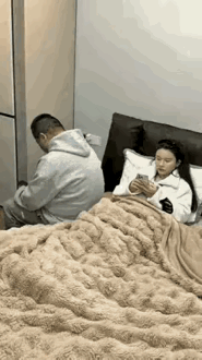老婆躺在床上看着手机，男士坐在床上突然接到父亲打来的电话站起身来，父亲告知他，母