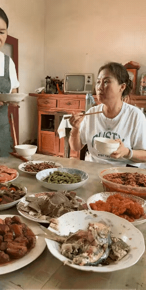 年轻女子刚端着菜走过来，还没来得及把菜盘子放到桌子上，坐在桌上的年长一点的女子就