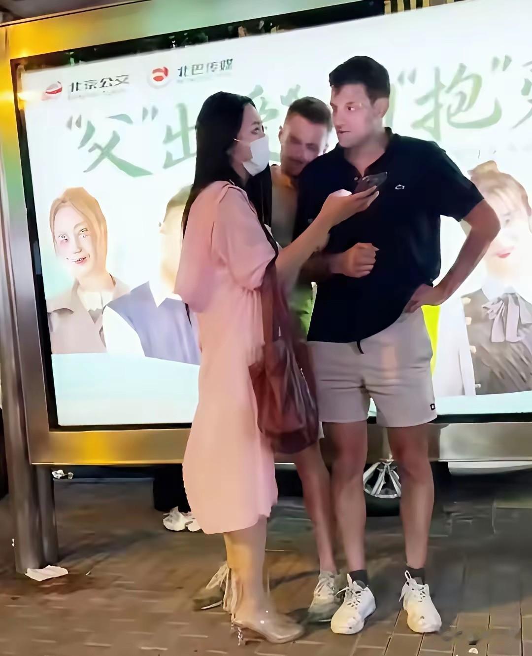 这个穿着粉红色连衣裙，脸上戴着口罩，手上拿着手机的美女，看到眼前的外国人，主动上