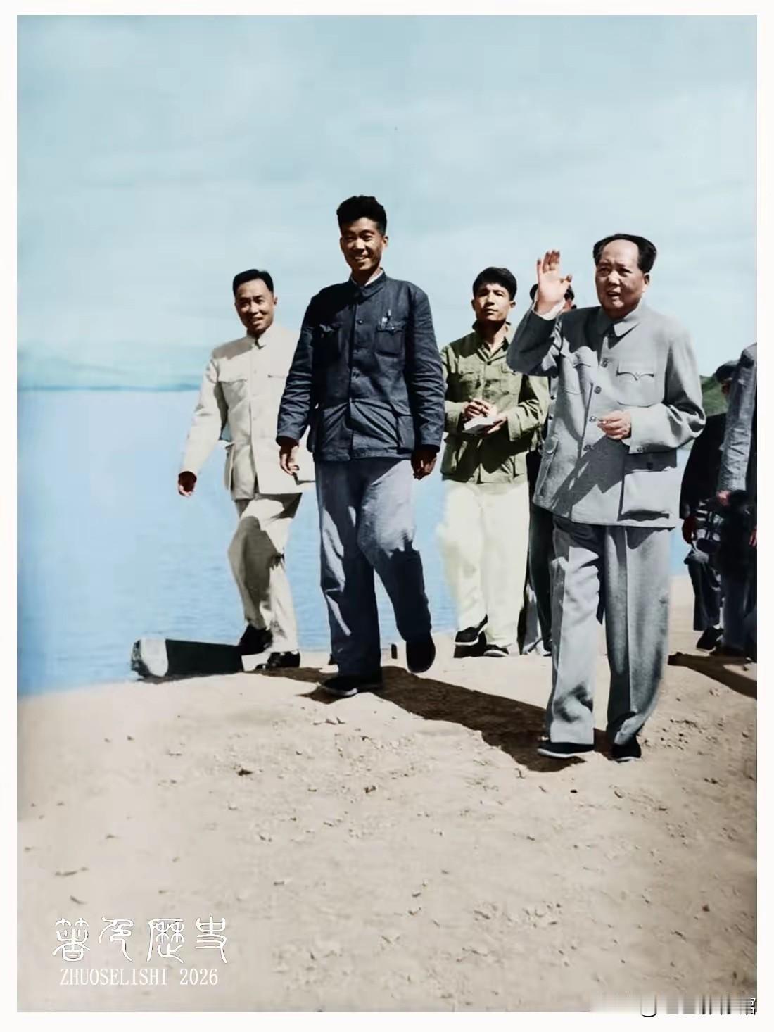 这张上色历史影像记录毛主席湖边视察的场景，他身着灰中山装抬手挥手，身旁随行干部缓