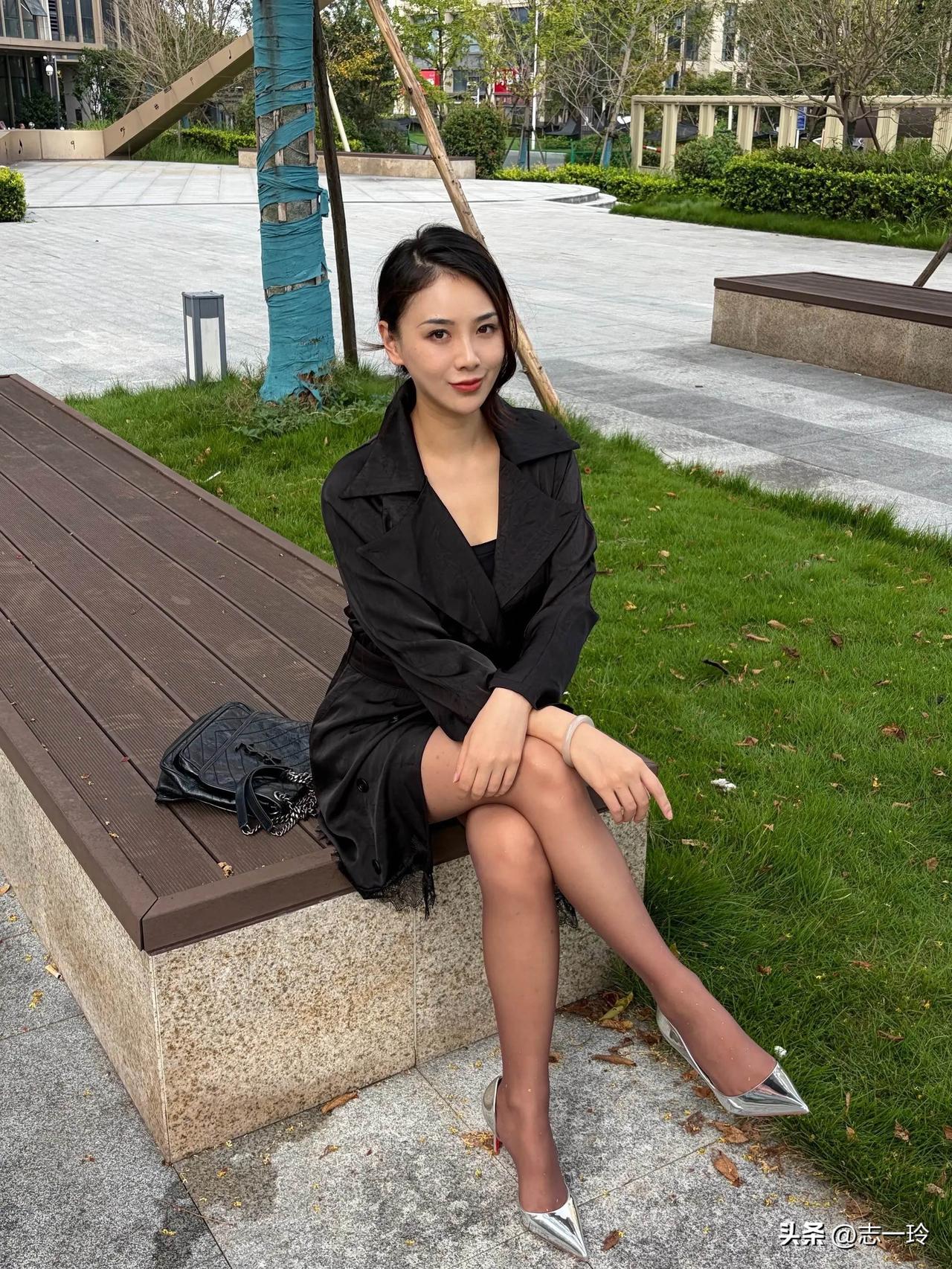 这位女士坐在公园长椅上，穿着黑色风衣和银色高跟鞋。