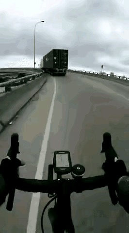 骑行者在公路转弯处想超过前方急驰的大货车，结果被大货车的尾部掀翻，所幸没有进入货