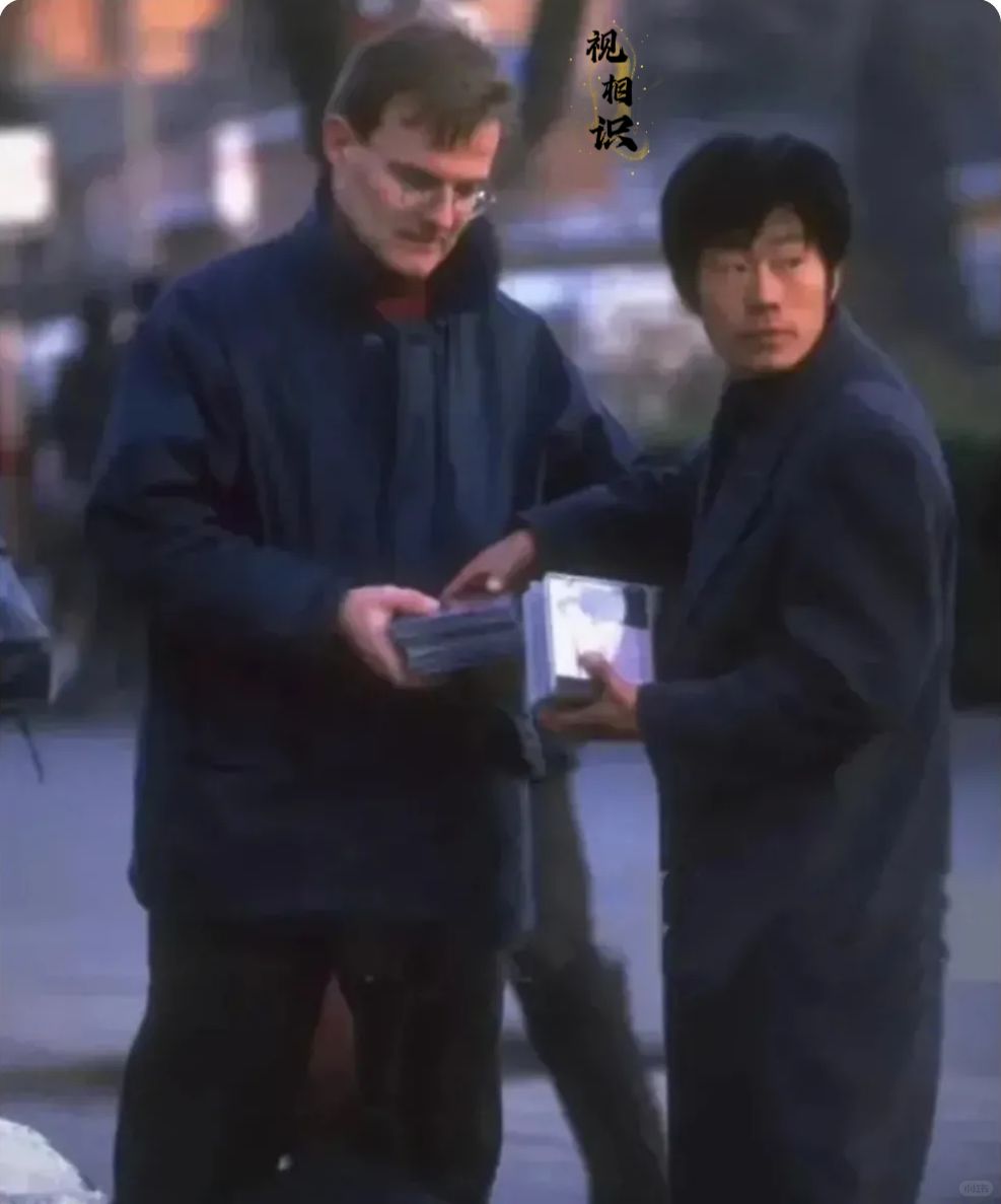 90年代，北京，一位男子正在向外国游客推销盗版光盘的旧照，画面中他还四处张望，显...