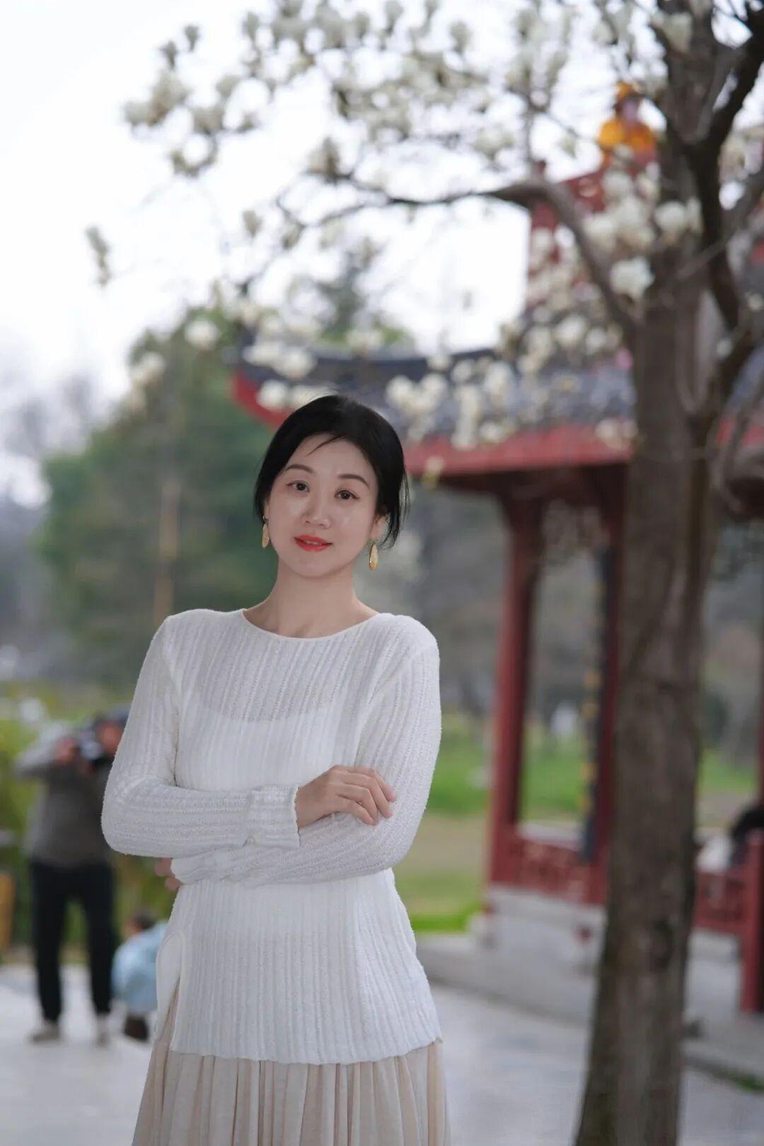 她置身于古色古香的中式园林中，背景是朱红廊柱、雕花栏杆与黛色飞檐，枝头盛放的白玉