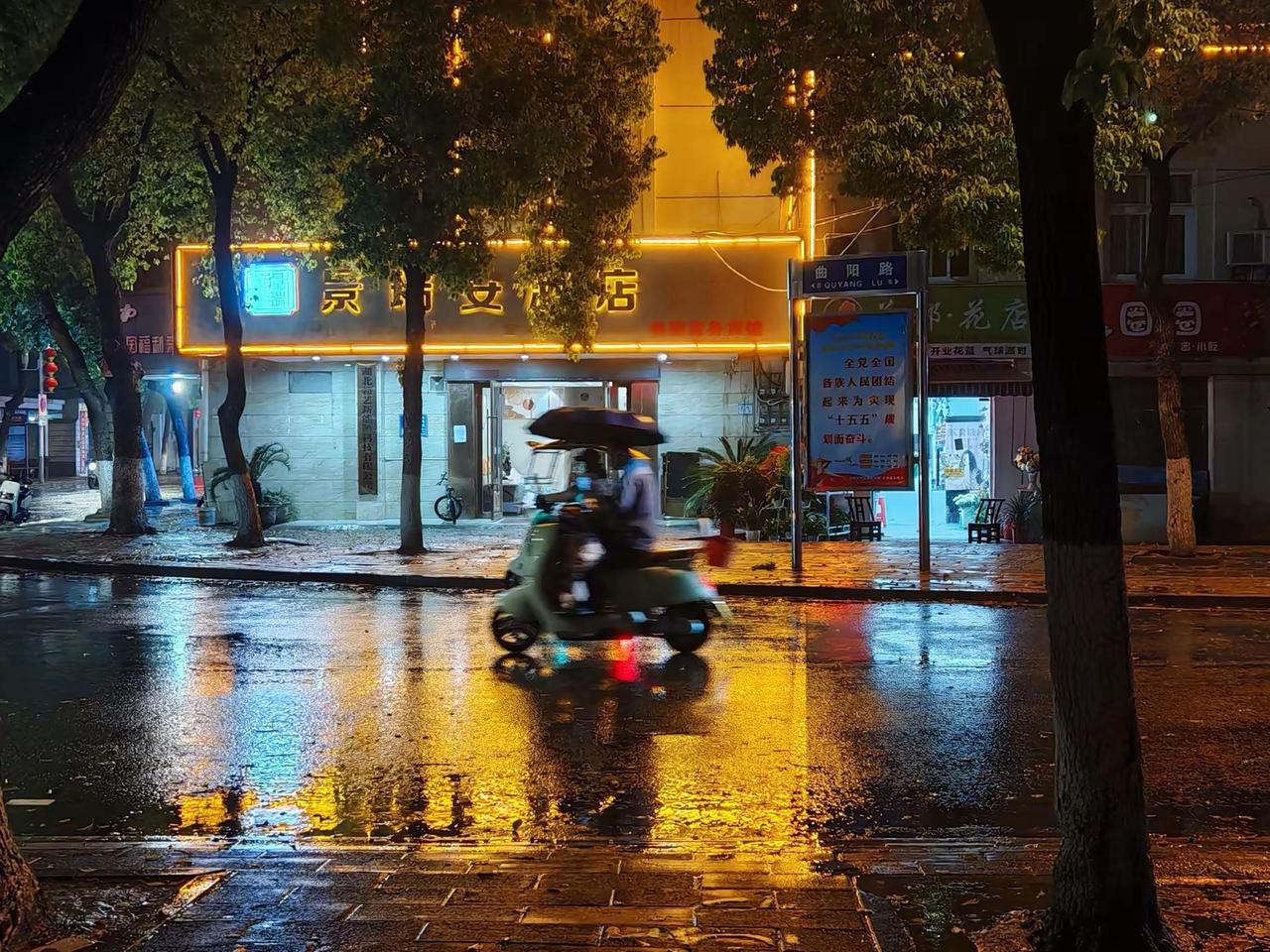 春雨夜。夜幕低垂，街灯在雨中朦胧成一片温暖的光晕，模糊了现实与梦境的边界。骑车人