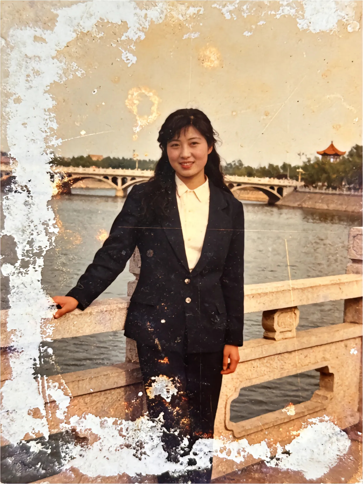 这是一张1988年的留影，照片中身着崭新西装的女子倚在河边石栏旁，笑容里满是那个