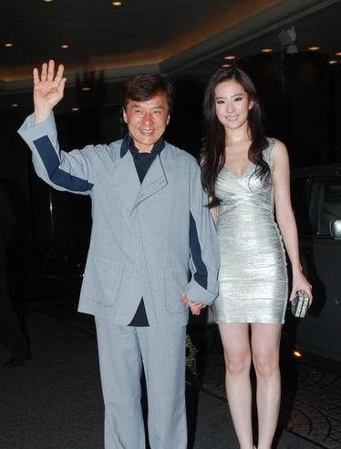 画面中身穿浅灰拼接休闲套装的成龙气色盈和，向镜头挥手致意；一旁身着银色短款礼服的