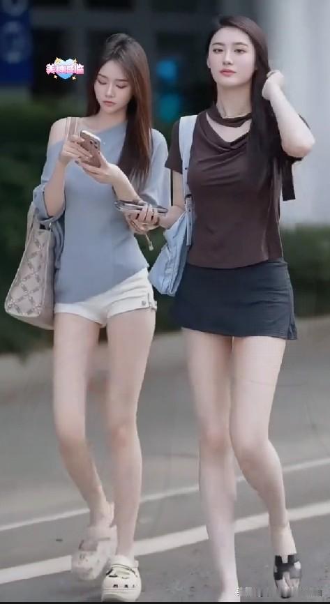 街头林荫步道上，两位结伴出行的女生穿搭清爽亮眼。左侧女生垂眸刷着手机，灰调露肩小