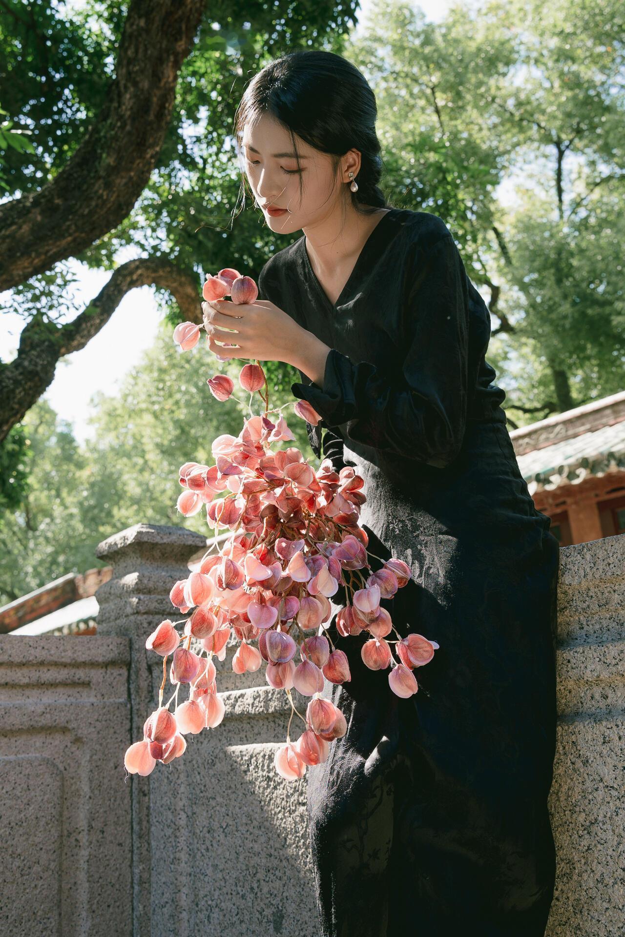 这组照片以黑色丝质长裙为基底，搭配粉调花束，在庭院场景中完成了“冷与暖”的视觉平
