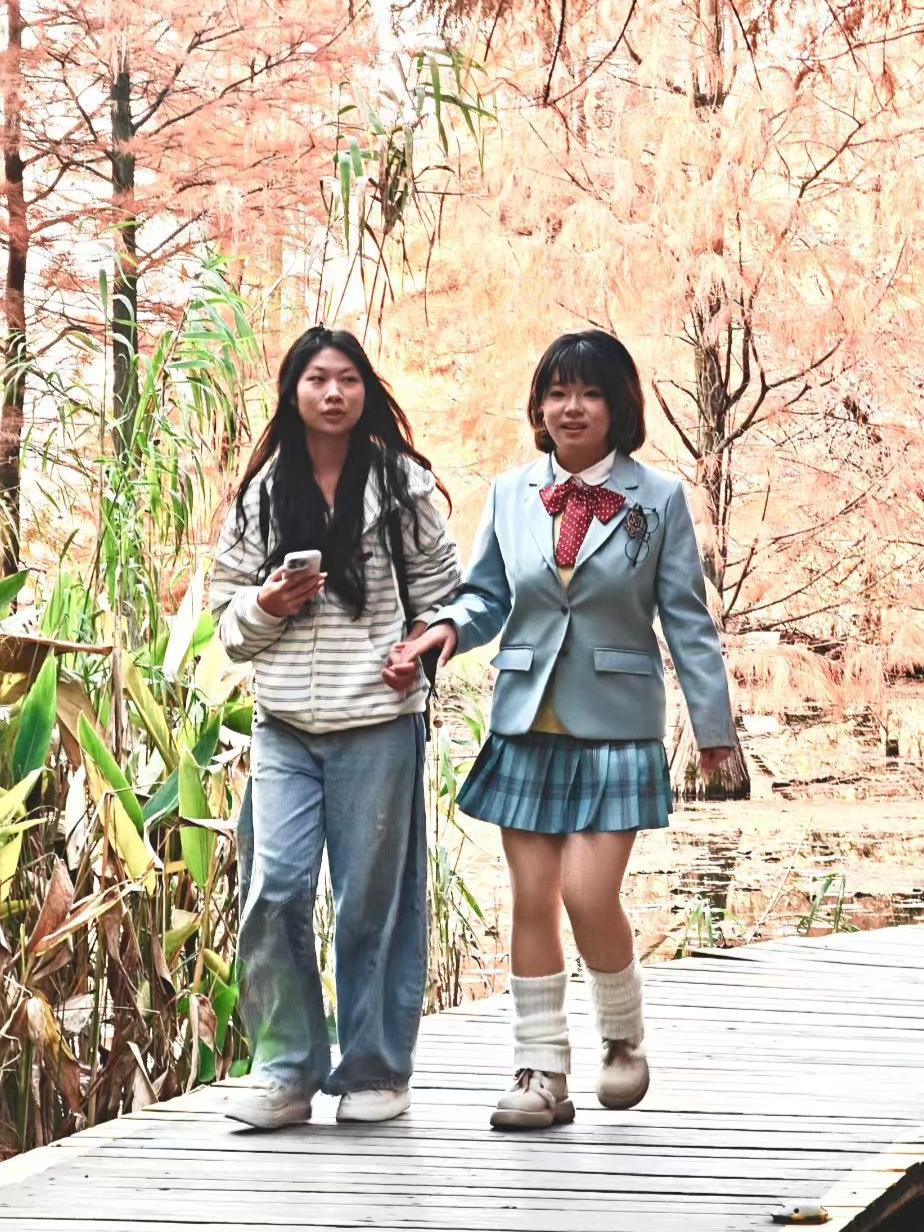 秋林把暖意揉进松针里，灰调大衣裹着轻软的风，红棕棉服倚着树影。她指尖亮着的屏幕，