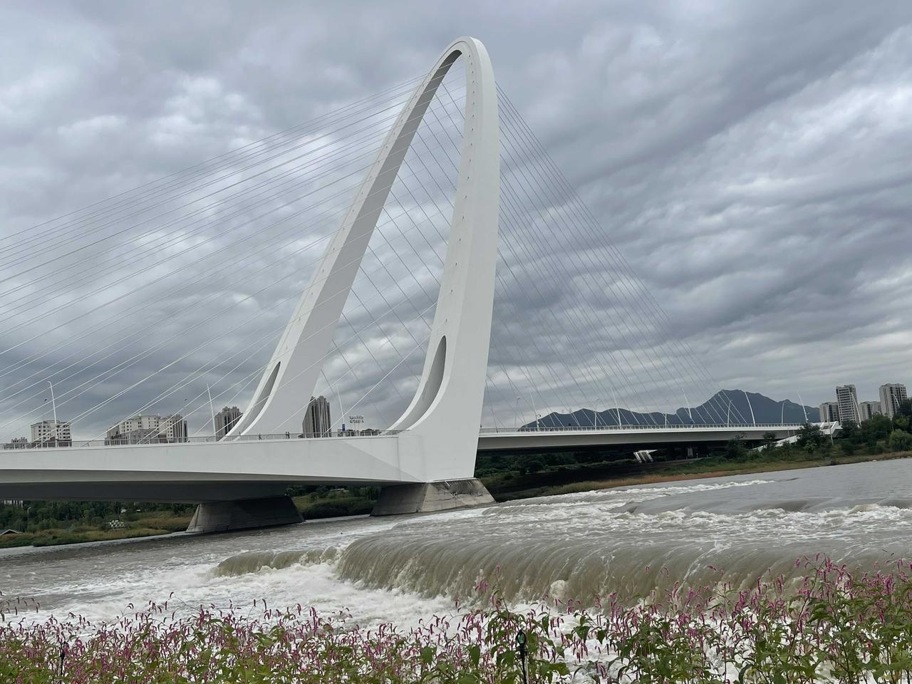 天空断续小雨，浓厚的云层下，新首钢大桥飞跨，永定河泛着波浪，石景山首钢园呈现油画