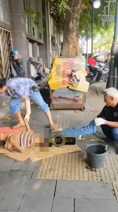 印尼的两名戴着手套的男子在城市人行道上，利用水桶和一些简单的用品，为一名躺在硬纸