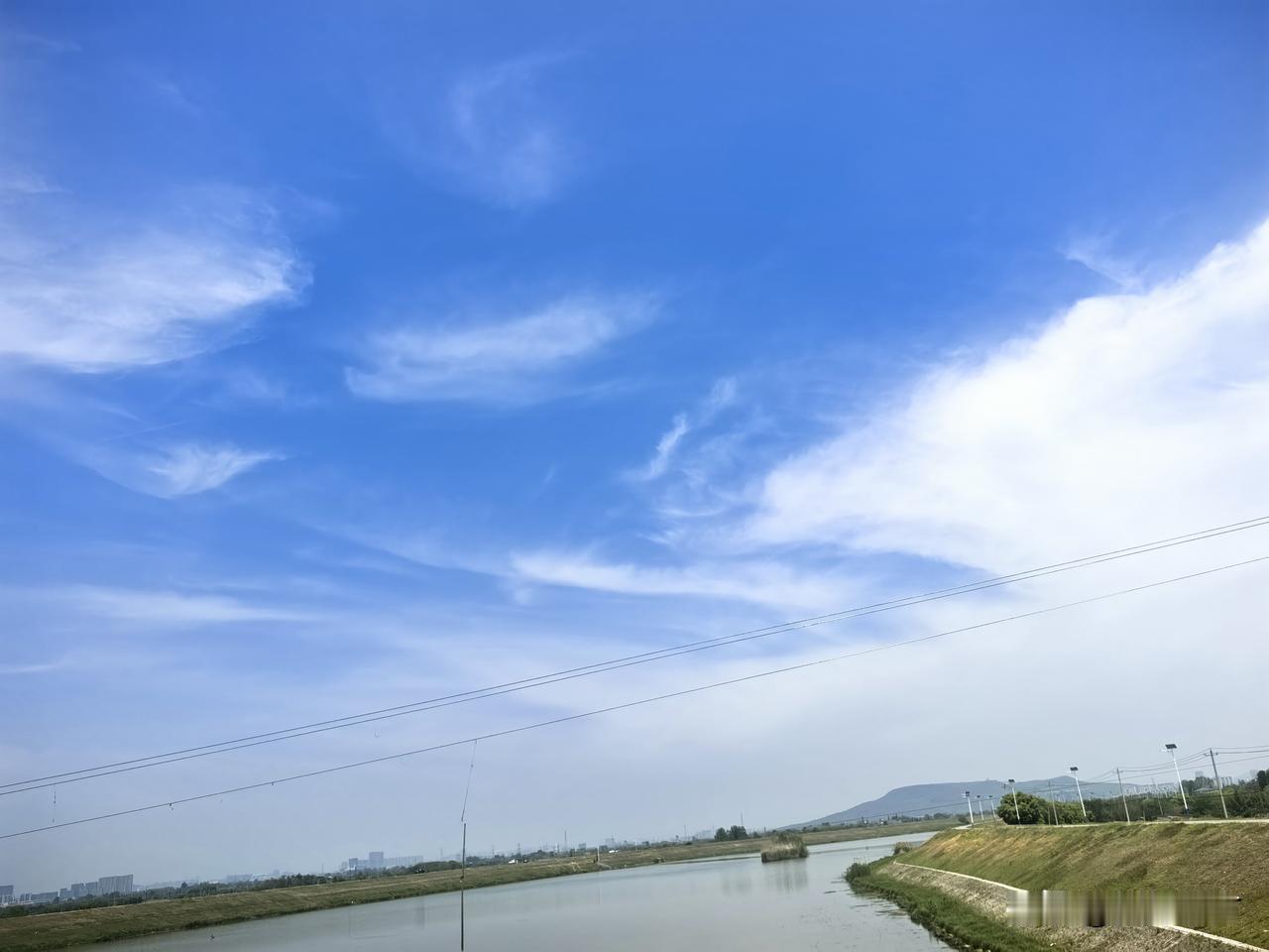 晴空如洗，流云舒展，河道蜿蜒向远，岸坡覆着新绿，远山淡影衬着城市轮廓，天地间清朗